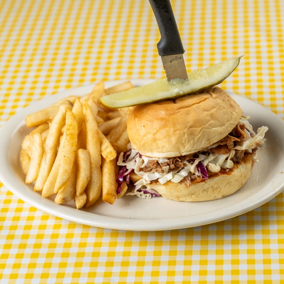 Pulled pork sandwich with coleslaw, pickle spear, and golden fries on a plate atop a yellow checkered tablecloth.