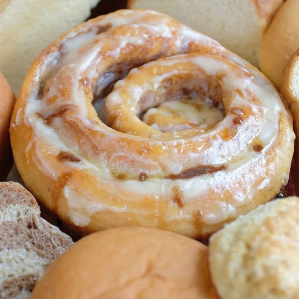 Glazed ring pastry surrounded by assorted bread rolls and slices of multigrain bread.