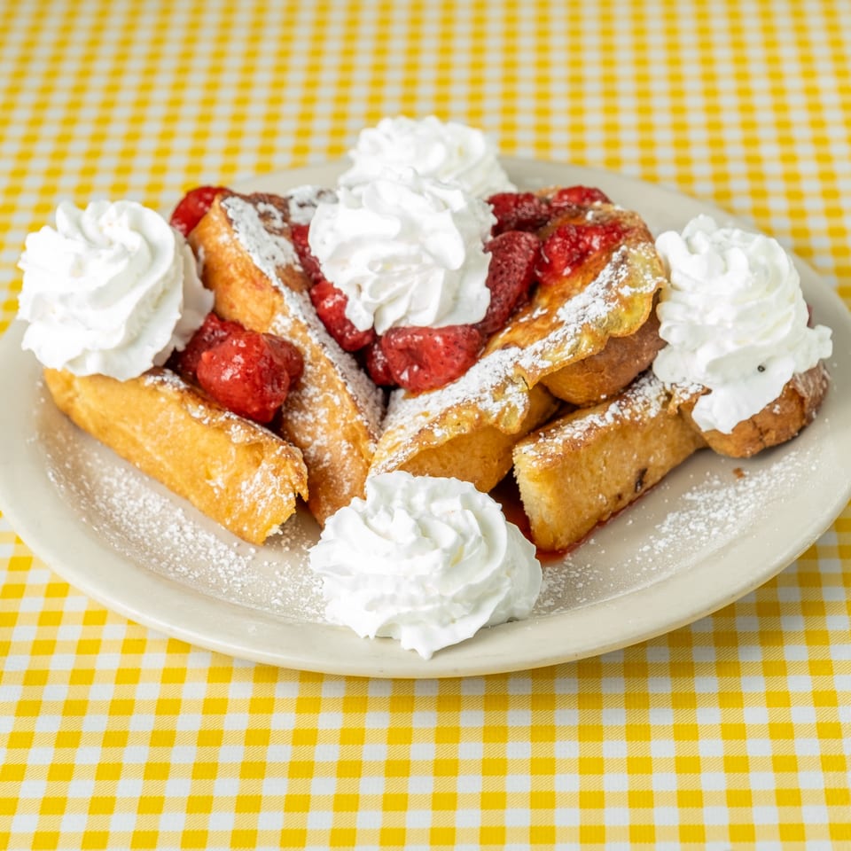 Plate of golden French toast topped with strawberries, powdered sugar, and whipped cream on a yellow checkered tablecloth.