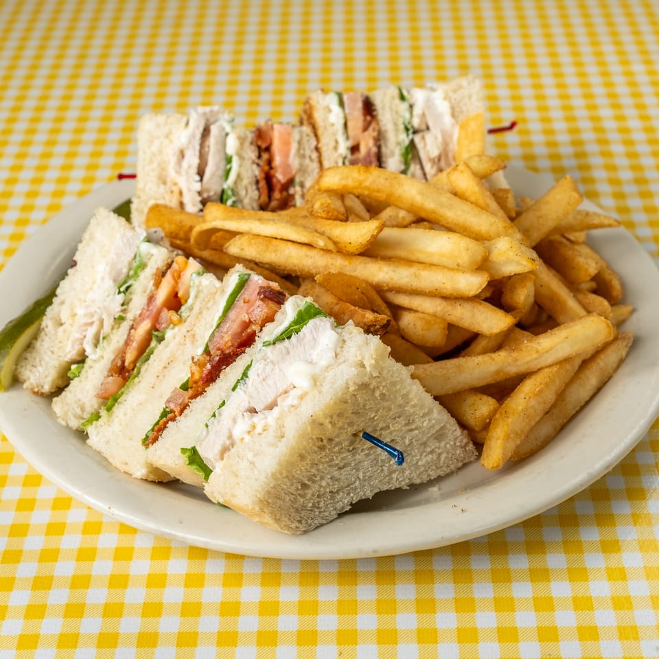 Plate of club sandwich quarters with lettuce, tomato, and bacon alongside golden fries and a pickle on yellow checkered tablecloth.