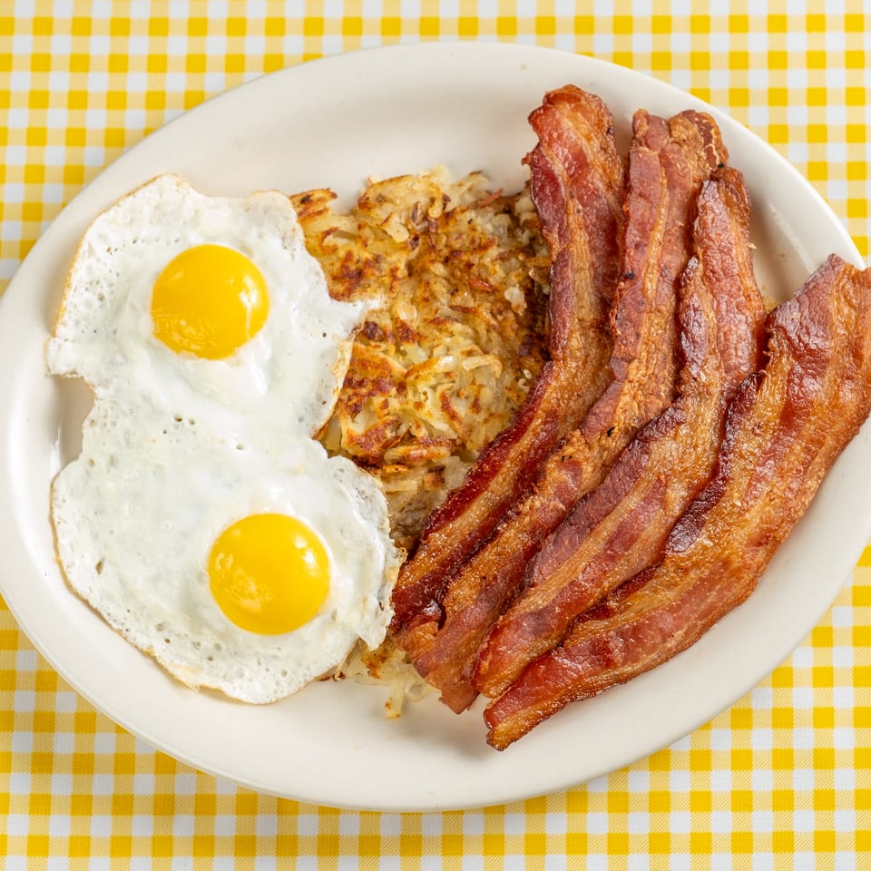 Plate of crispy bacon, two fried eggs, and golden hash browns on a yellow checkered tablecloth.