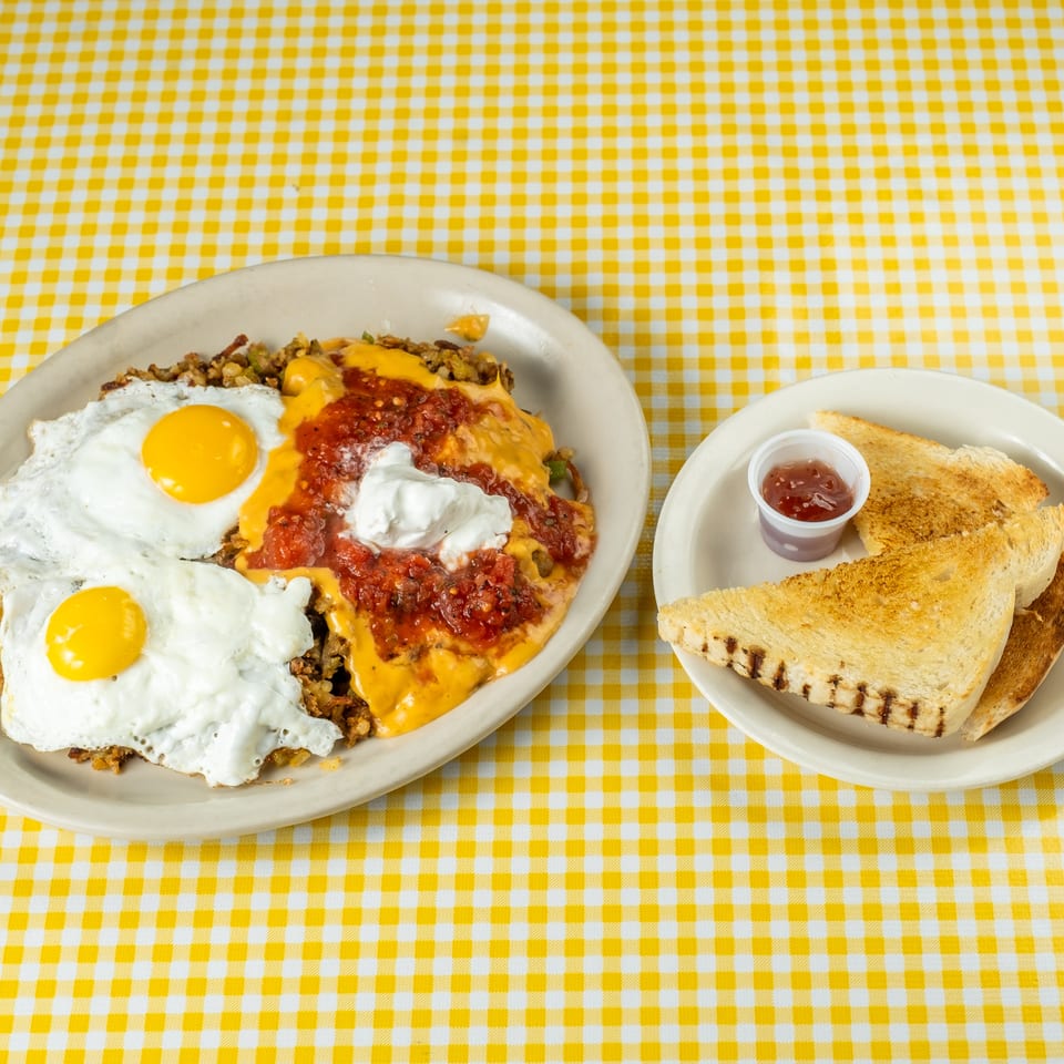 Breakfast plate with two fried eggs, cheesy hash browns, salsa, and sour cream beside grilled toast and jam on a yellow checkered tablecloth.