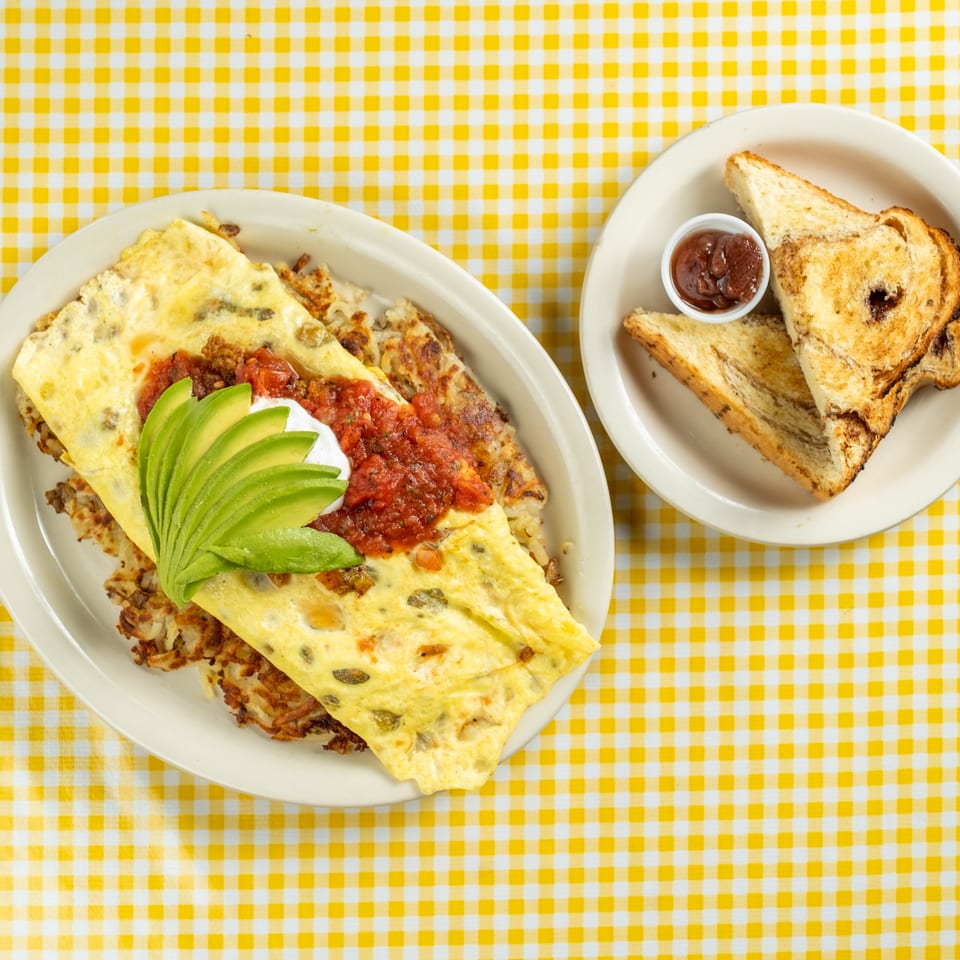 Cheesy omelet topped with salsa, avocado, and sour cream alongside toasted bread and jam on a yellow checkered tablecloth.