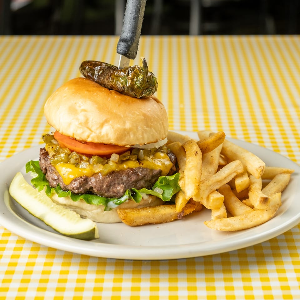Cheeseburger topped with grilled green chile, tomato, lettuce, and onions, served with fries and a pickle on a checkered tablecloth.