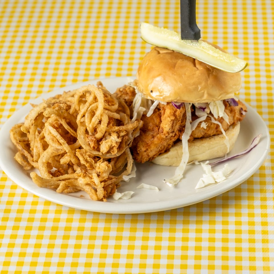 Crispy fried chicken sandwich with cabbage slaw, pickle garnish, and onion rings on a plate atop a yellow checkered tablecloth.