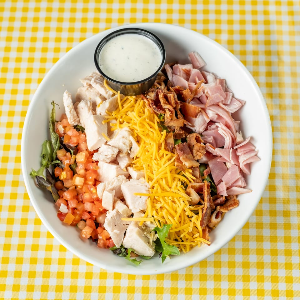 Chopped salad topped with grilled chicken, ham, bacon, shredded cheddar, tomatoes, greens, and ranch dressing on a checkered tablecloth.