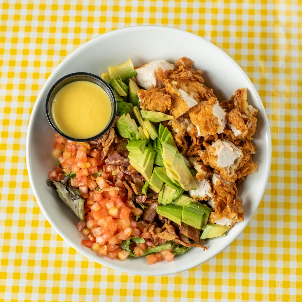 Crispy fried chicken salad with avocado, diced tomatoes, bacon, greens, and creamy dressing in a white bowl on yellow gingham.