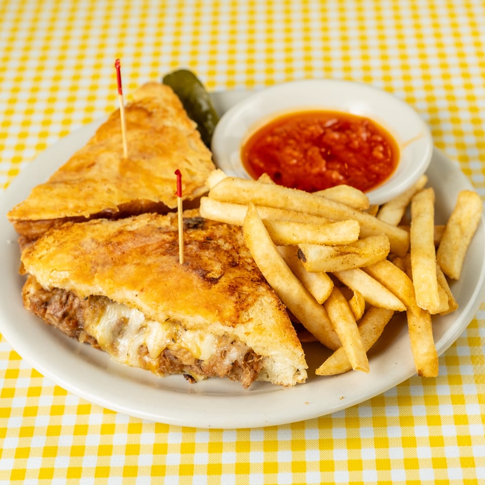Grilled cheese sandwich and triangular toasted sandwich served with French fries, marinara sauce, and pickle on a yellow checkered tablecloth.