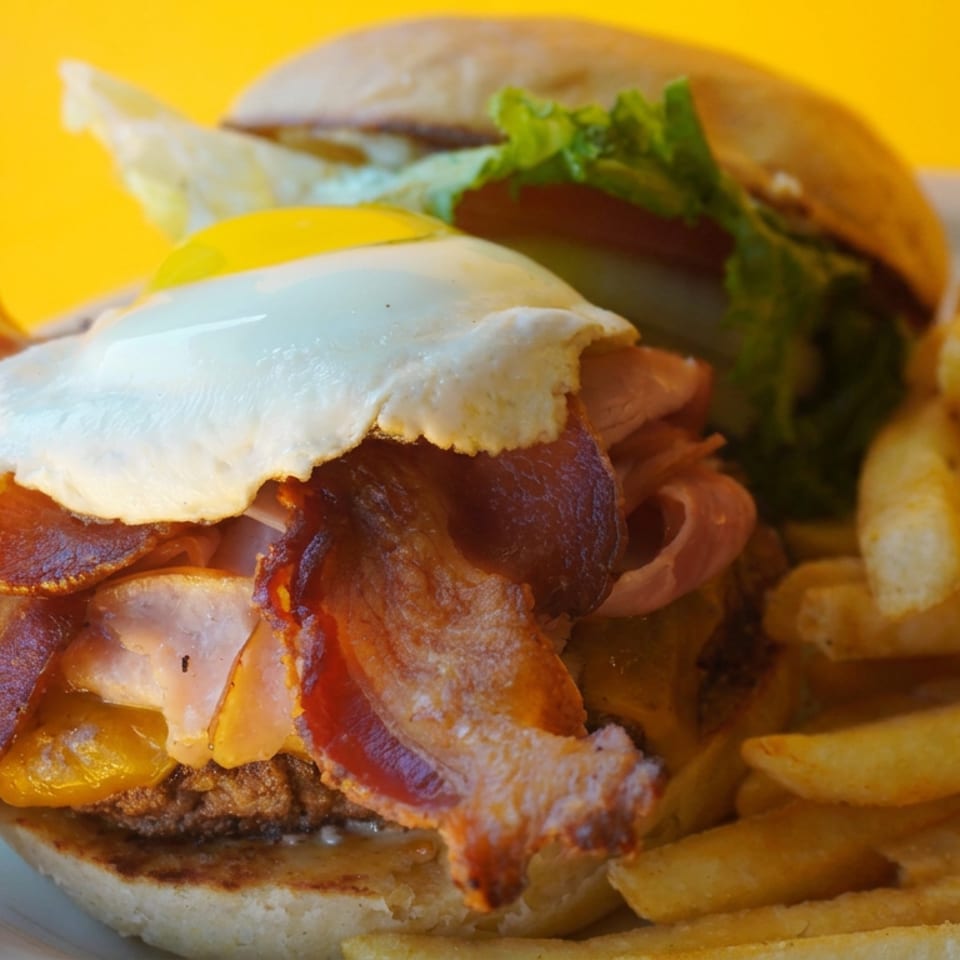 Bacon cheeseburger topped with a fried egg and vegetables, served with golden fries on a plate.