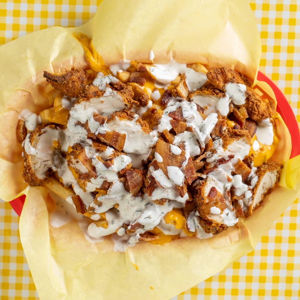 Loaded fries topped with fried chicken, bacon, cheese, and creamy ranch dressing on a yellow checkered tablecloth.