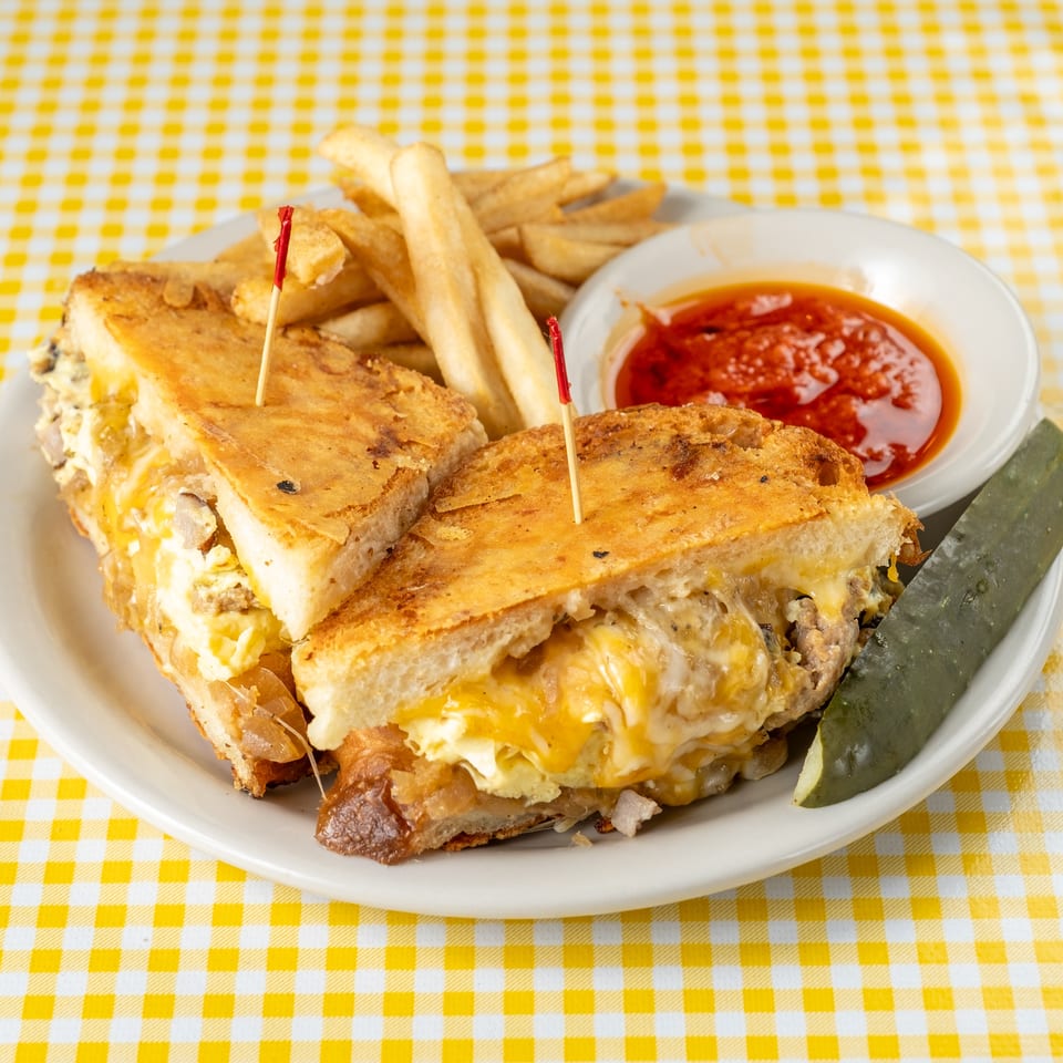Grilled cheese sandwiches with melted cheese, fries, ketchup, and pickle spears on a white plate atop a yellow checkered tablecloth.
