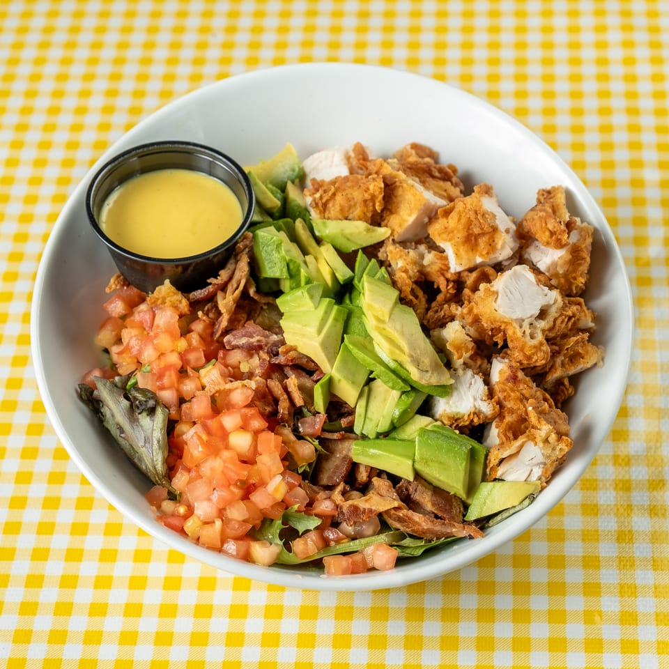 Loaded salad bowl with crispy fried chicken, avocado, bacon, tomatoes, greens, and dipping sauce on a yellow checkered tablecloth.