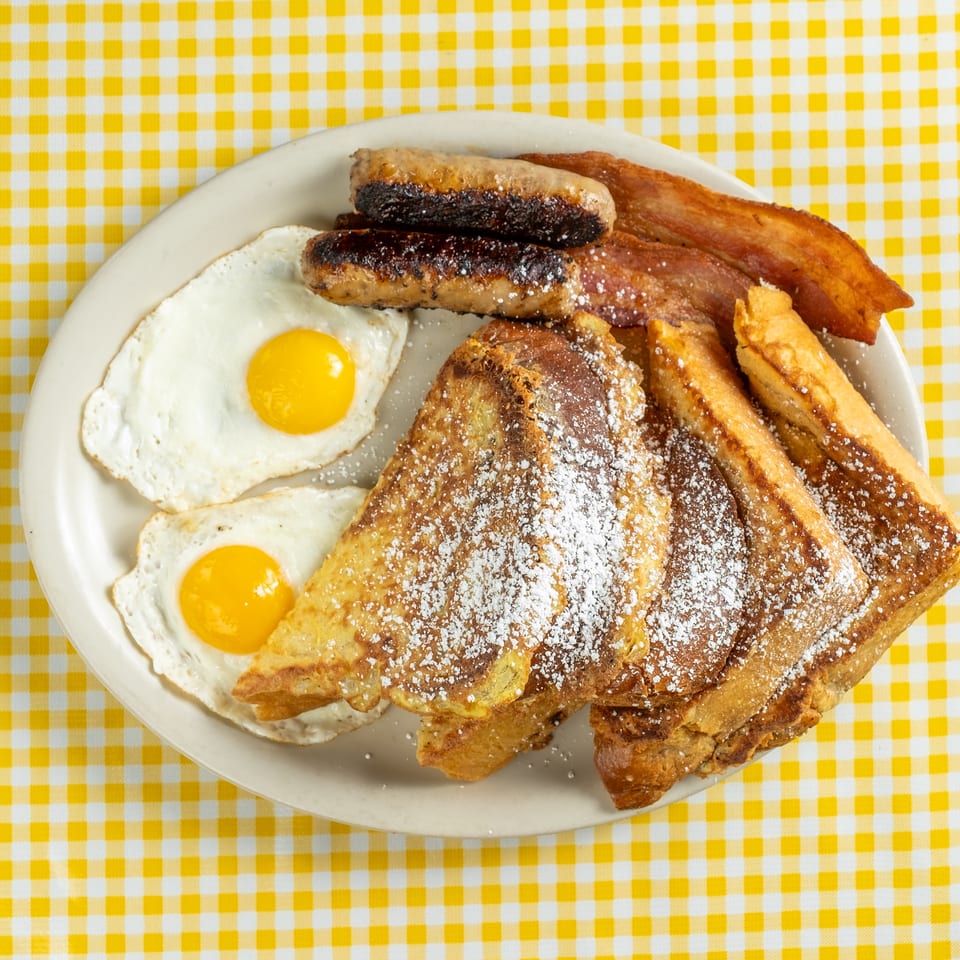 Breakfast plate with fried eggs, sausage, bacon, French toast dusted with powdered sugar, and home fries on yellow gingham cloth.