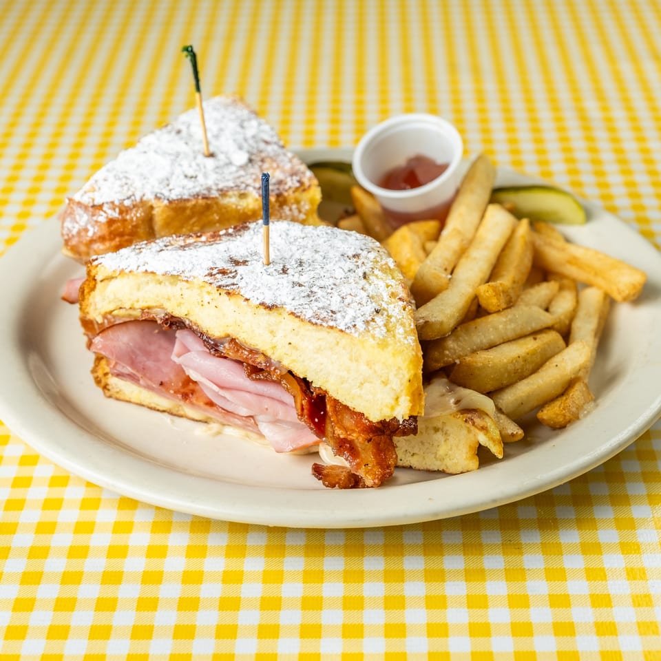 Powdered-sugar-dusted ham and bacon sandwich served with fries, pickles, and ketchup on a yellow checkered tablecloth.