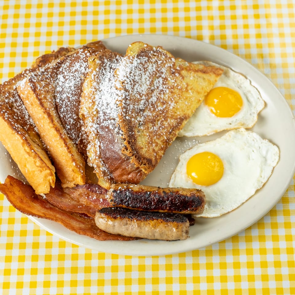 Breakfast platter with powdered-sugar French toast, fried eggs, bacon, and sausage on a yellow checkered tablecloth.