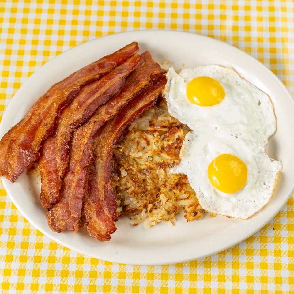 Plate of crispy bacon, two sunny-side-up eggs, and golden hash browns on a yellow checkered tablecloth.
