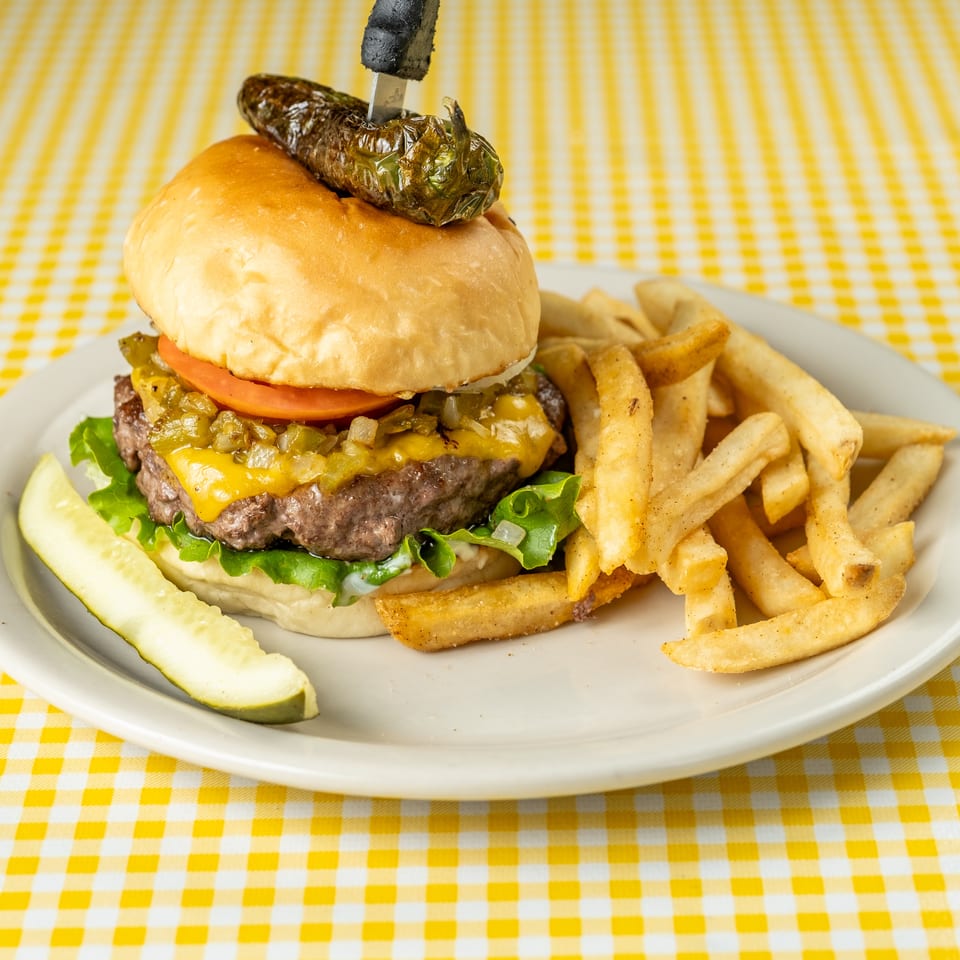Cheeseburger topped with tomato, lettuce, relish, and a grilled jalapeño, served with fries and a pickle spear on a checkered tablecloth.