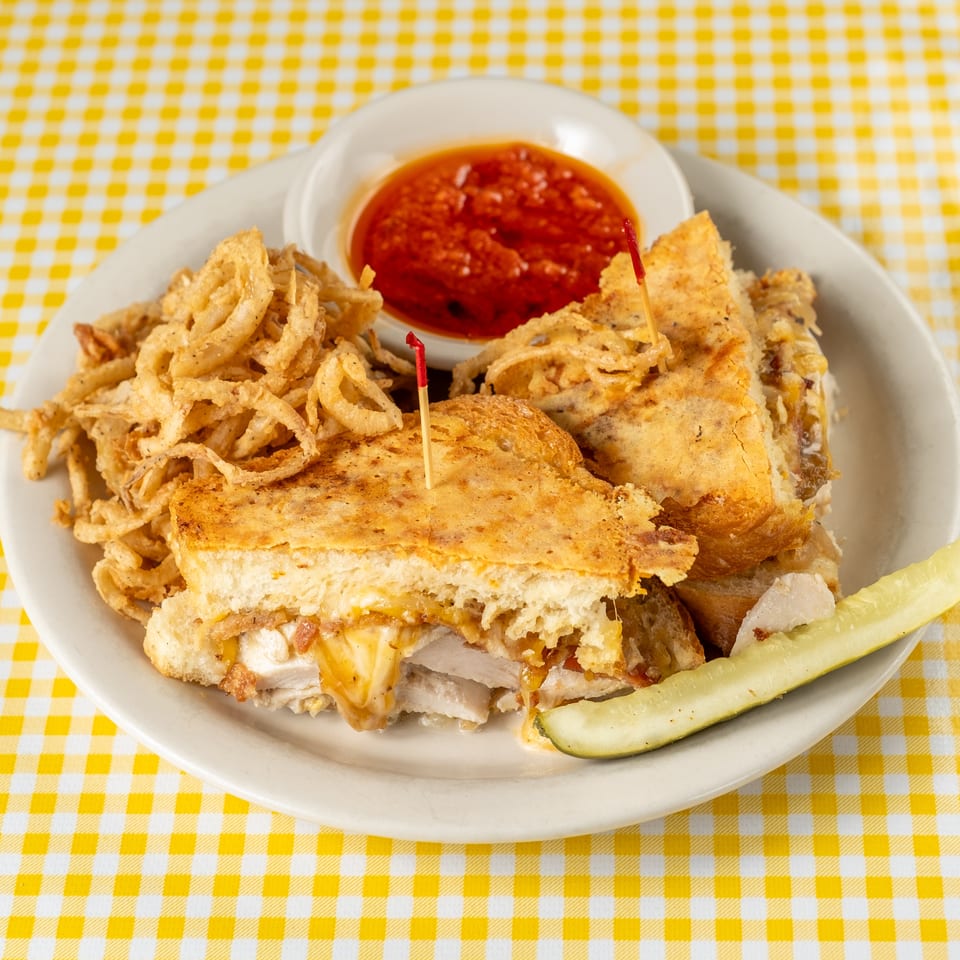 Grilled sandwich with melted cheese, crispy onion rings, red dipping sauce, and pickle spear on a white plate.