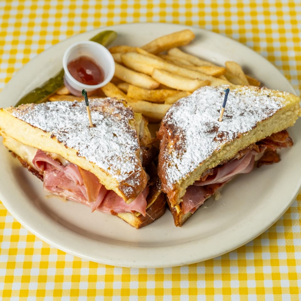 Powdered-sugar-dusted ham and cheese sandwiches with fries, ketchup, and pickle on a plate atop a yellow checkered tablecloth.