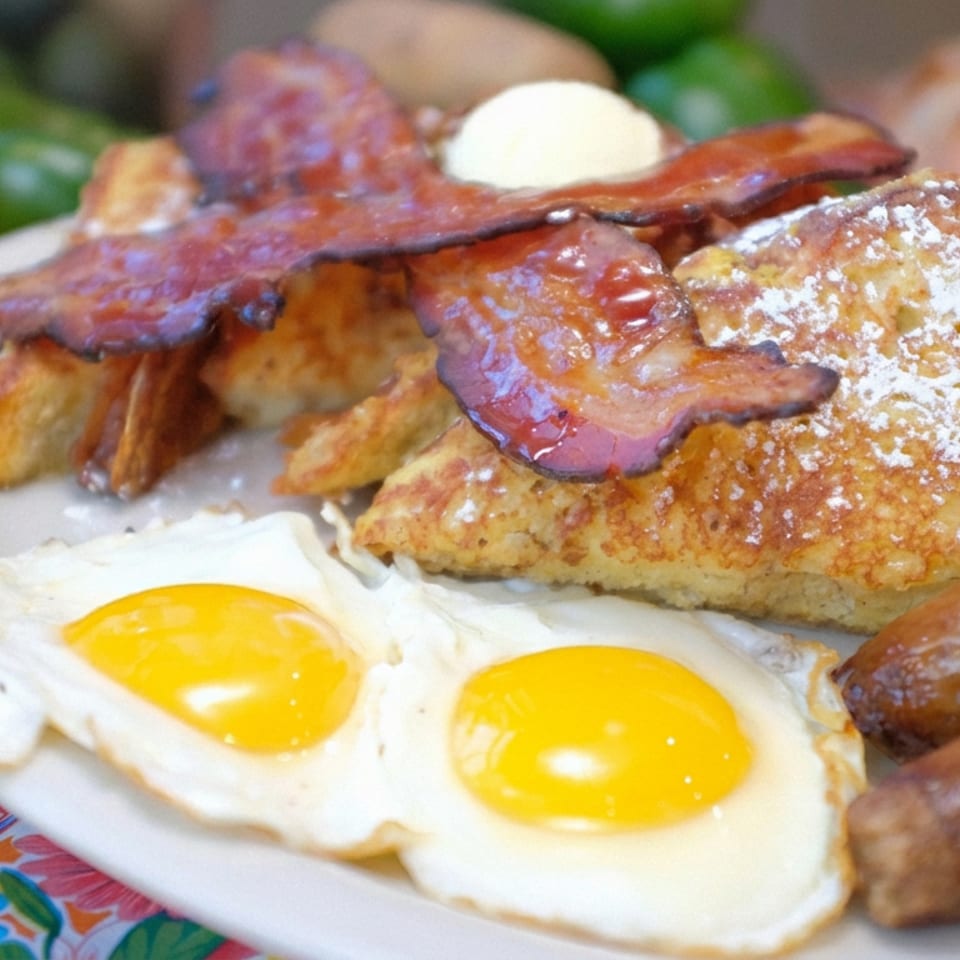Brunch plate with two fried eggs, sausages, home fries, bacon, powdered-sugar French toast, and butter.