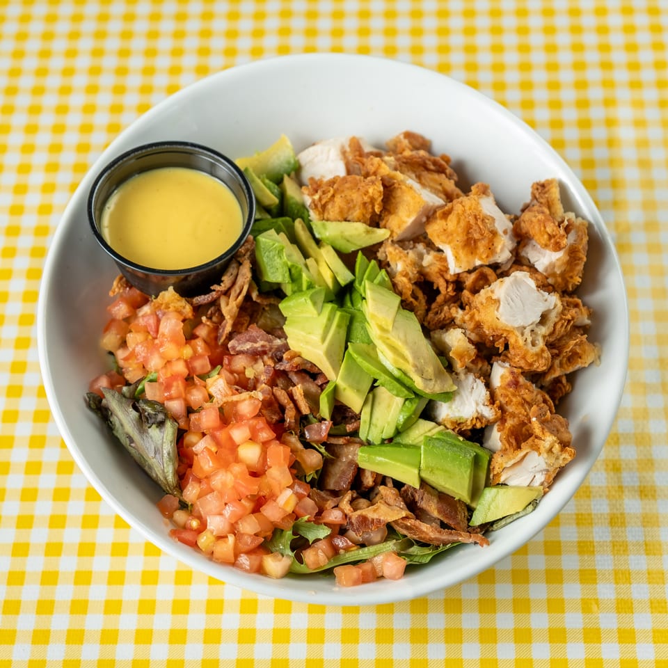 Cobb salad topped with crispy fried chicken, avocado, tomatoes, bacon, greens, and dressing on a yellow checkered tablecloth.